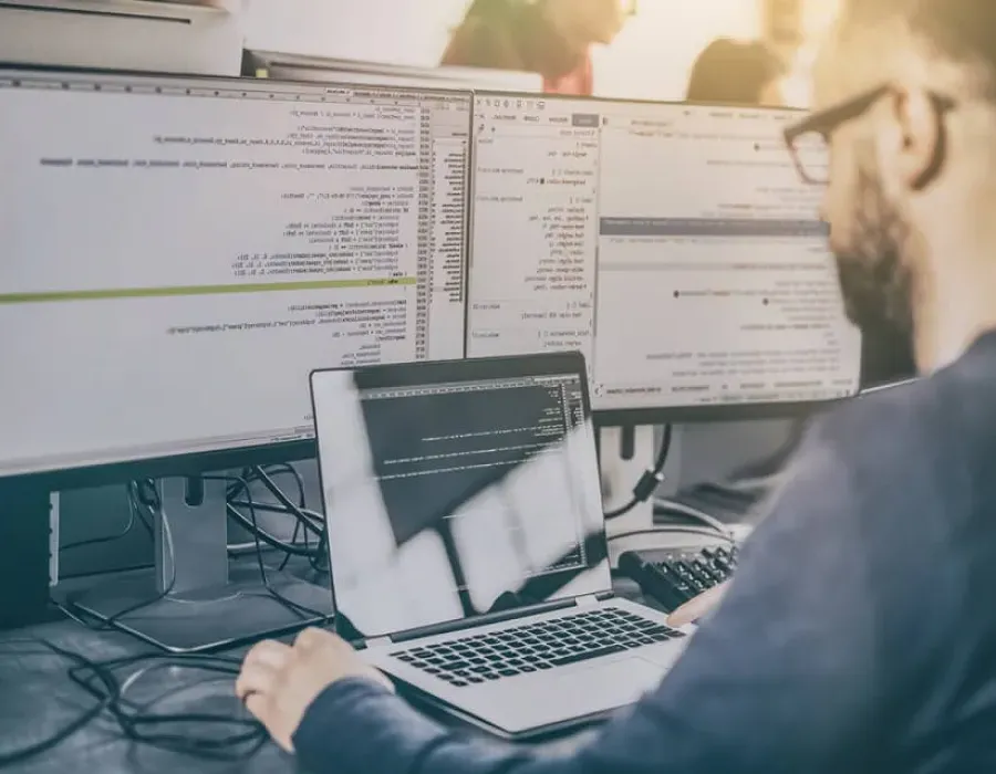 Computer programmer seated in front of two monitors and laptop executing code